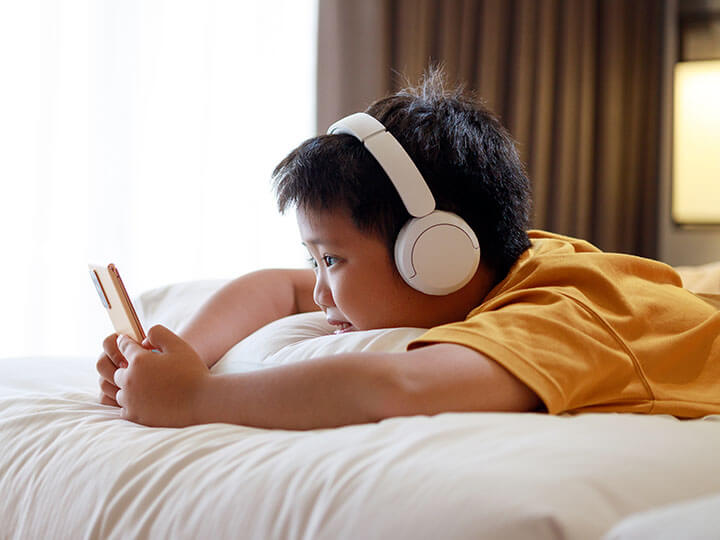 Asian child lying on a bed face down looking at a smartphone while listening through white wireless headphones