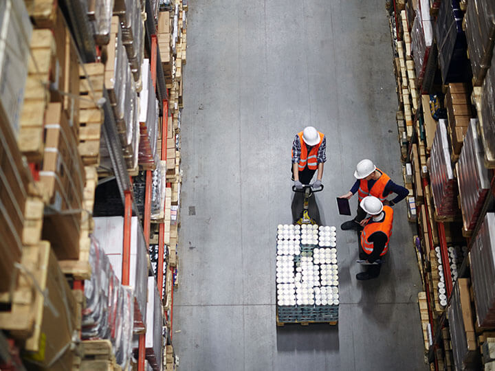 Group of workers in construction caution vests and hardhat helmets having talk while looking at a table in a warehouse aisle