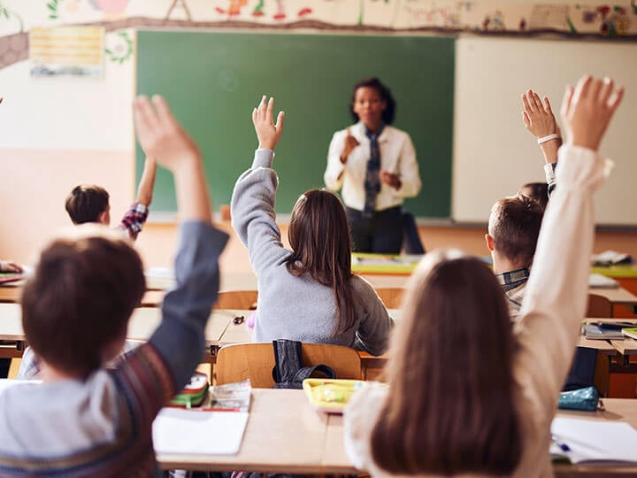 Rear view of students raising their hands in classroom as an African American female teacher asked a question in front of a green chalk board