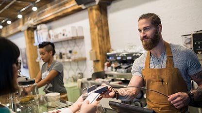 Woman paying for a beverage in a coffee shop, exchanging payment with a smiling, bearded, young man.