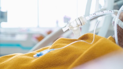 A patient in a hospital bed with a yellow blanket is connected to a breathing tube and an IV line.